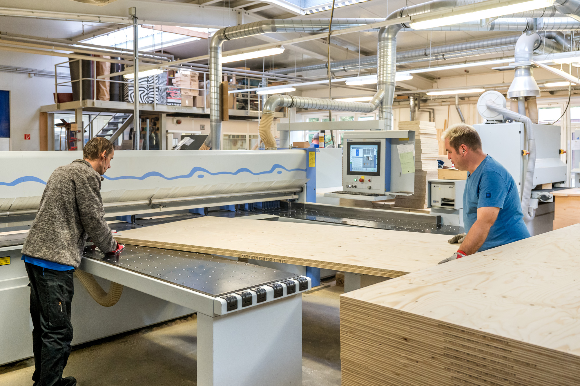 Zwei Mitarbeitende der Werkstatt verarbeiten an einer hochmodernen Plattensäge ein große Holzplatte