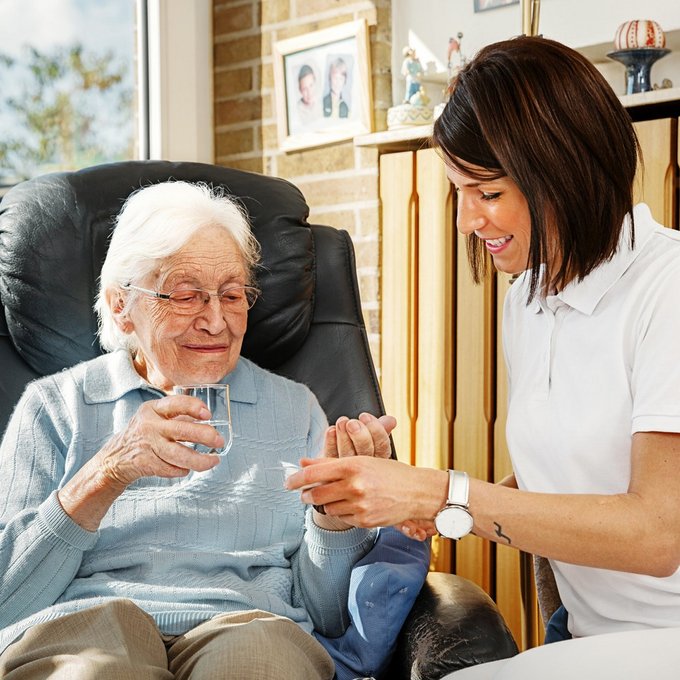 Eine Seniorin sitzt zuhause in einem gemütlichen schwarzen Ledersessel vor dem Fenster. Sie bekommt von einer Pflegefachfrau ihre Medikamente gereicht und hält ein Glas Wasser in Ihrer rechten Hand