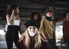 Ein Mädchen sitzt am Boden und macht eine Kaugummiblase. Sie ist von anderen Jugendlichen umgeben