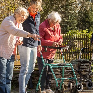 drei Senioren mit Rollator und eine Begleiterin beim Spazieren im Grünen