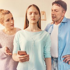 Eine Teenagerin steht mit Kopfhörern und geschlossenen Augen zwischen ihren Eltern. Die Eltern reden auf sie ein. 