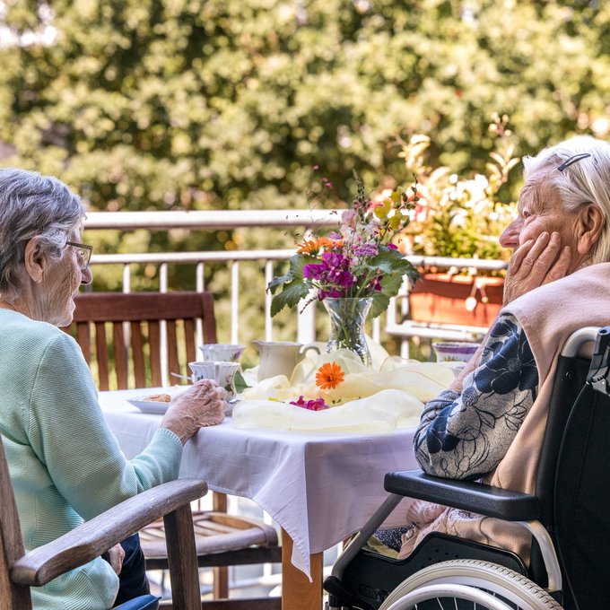 Zwei ältere Damen, eine im Rollstuhl, sitzen bei Sonnenschein auf der Terrasse, schauen ins Grüne und trinken gemeinsam Kaffee