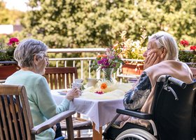 Zwei ältere Damen, eine im Rollstuhl, sitzen bei Sonnenschein auf der Terrasse, schauen ins Grüne und trinken gemeinsam Kaffee