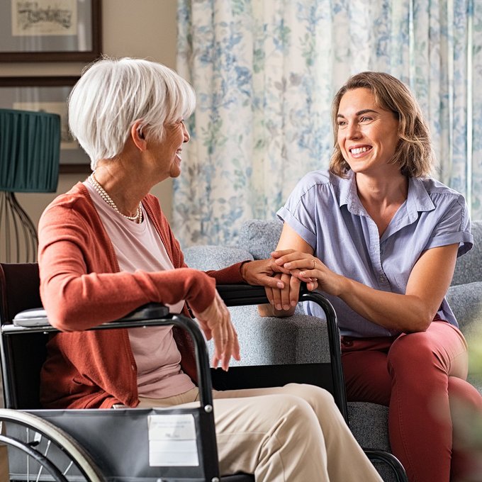 Eine Seniorin im Rollstuhl sitzt mit Ihrer Tochter im Wohnzimmer, beide lächeln sich an und die Tochter hält eine Hand der Mutter in beiden Händen.