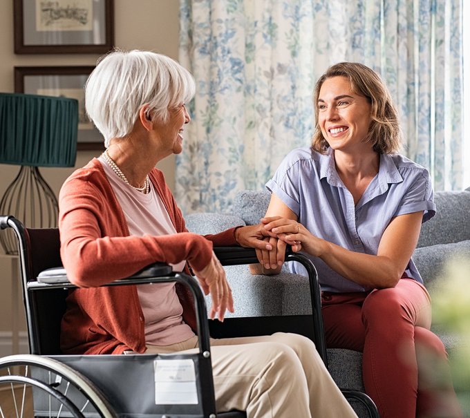 Eine Seniorin im Rollstuhl sitzt mit Ihrer Tochter im Wohnzimmer, beide lächeln sich an und die Tochter hält eine Hand der Mutter in beiden Händen.