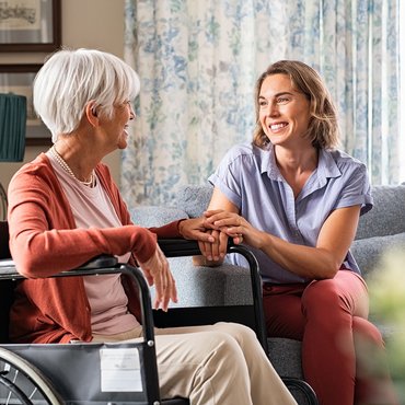 Eine Seniorin im Rollstuhl sitzt mit Ihrer Tochter im Wohnzimmer, beide lächeln sich an und die Tochter hält eine Hand der Mutter in beiden Händen.