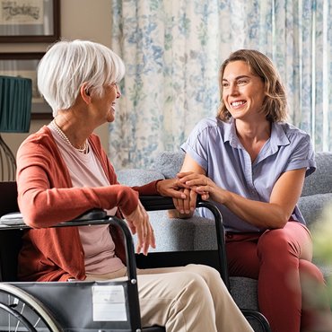Eine Seniorin im Rollstuhl sitzt mit Ihrer Tochter im Wohnzimmer, beide lächeln sich an und die Tochter hält eine Hand der Mutter in beiden Händen.