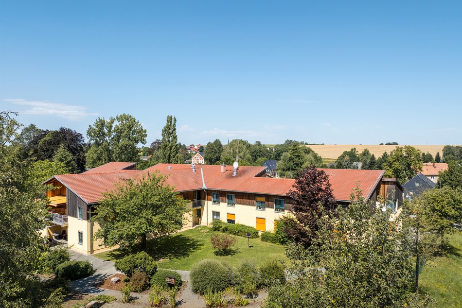 Luftbildaufnahme vom Haus Elisabeth vor strahlend blauem Himmel, das Gebäude hat hellgelbe Wänden und Dach mit roten Dachziegeln, es steht in Mitten von Bäumen